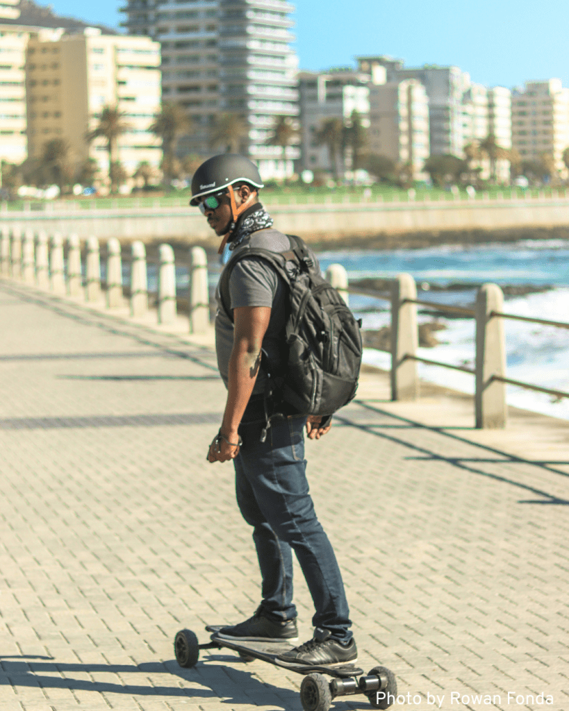 An older man rides an electric skateboard along a beachside town, enjoying the sunny day and coastal scenery.
