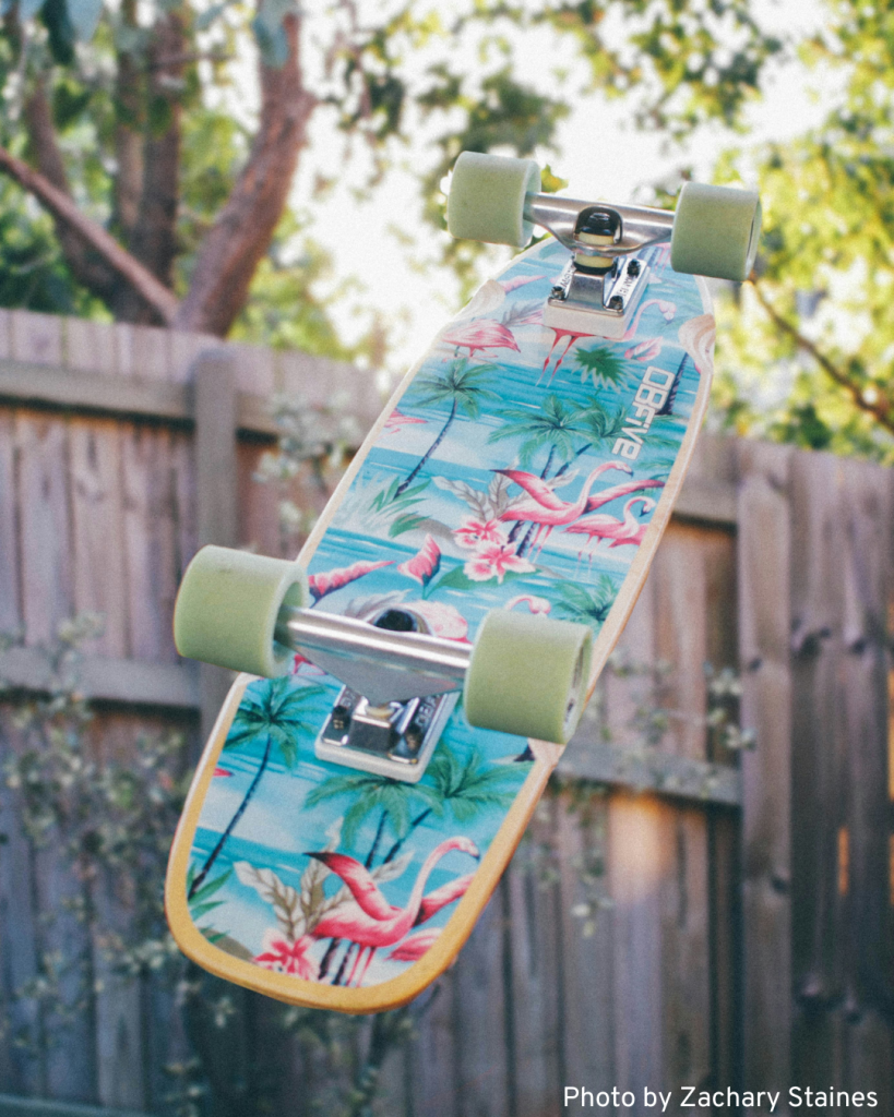 A tropical print cruiser skateboard hangs from a fence, showcasing vibrant colors and a fun design.