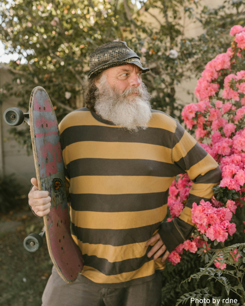 A bearded man poses with an 80's old-school skateboard, reflecting a nostalgic skateboarding spirit.